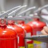 Feuerlöscher aufgereiht, man sieht eine Hand, die auf einem Klemmbrett etwas einträgt